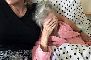 An elderly woman in a pink robe sits on a bed, covering her face with her hand. Beside her, another woman sits close, offering support. The bed is covered with a polka-dot blanket.